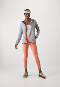 Svetlo siva jakna z zadrgo in črnimi poudarki, bela majica brez rokavov in svetlo oranžne pajkice. Nosijo zelene čevlje s črnimi in oranžnimi podplati.