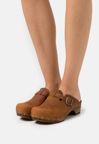 Zoccoli in pelle marrone con borchie metalliche e cinghie regolabili, indossati a piedi nudi, mostrati su uno sfondo bianco semplice.