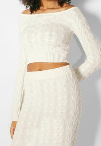 White cable knit crop top with off-the-shoulder design and ribbed hem, paired with a matching skirt featuring similar knit patterns.