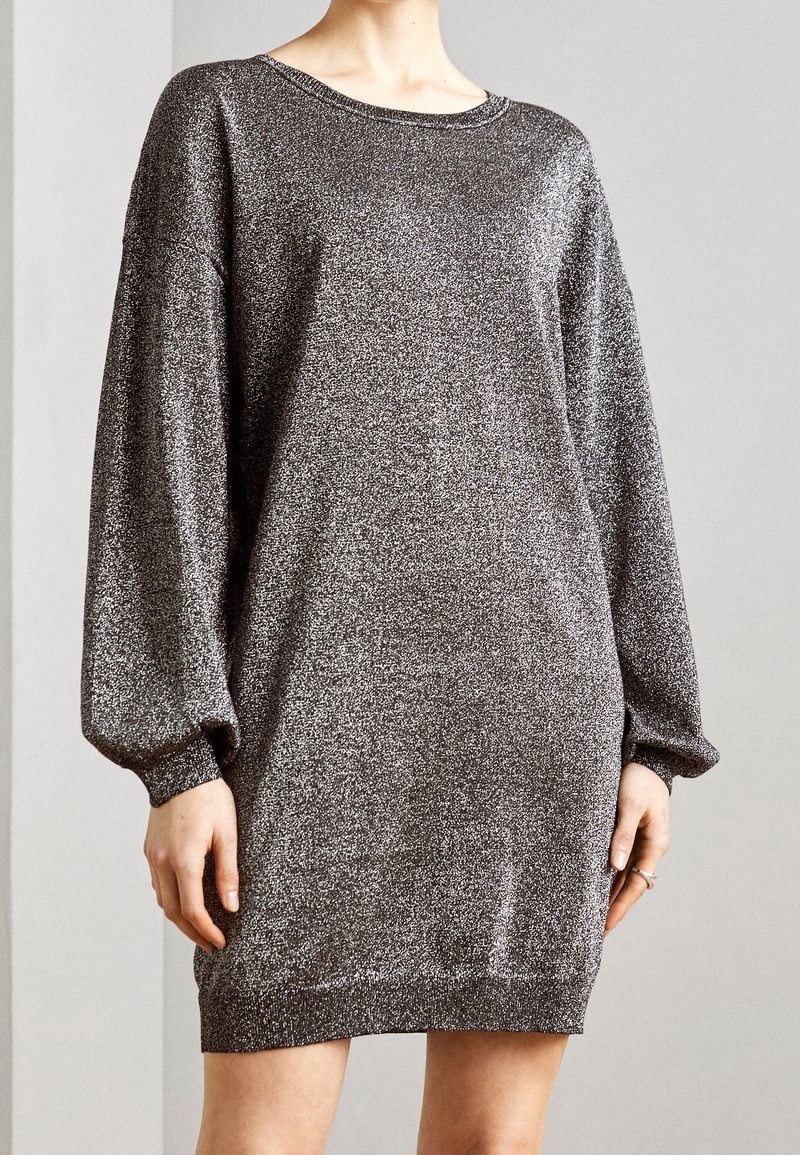 Robe en maille argentée scintillante à manches longues et longueur genou avec un col rond portée par une personne debout sur un fond uni.