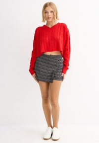 Suéter corto de punto rojo con diseño de trenzas, combinado con una falda corta de cuadros gris con botones y detalle lateral abierto. Zapatillas blancas.
