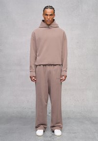 Felpa beige con tasca frontale e cappuccio regolabile, abbinata a pantaloni della tuta beige coordinati. Presenta dettagli ricamati sottili e sneakers bianche.