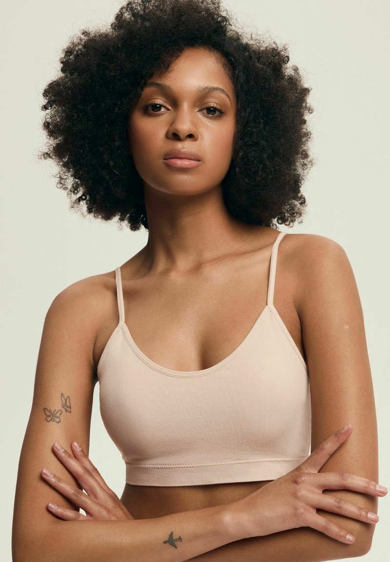 Jasnoróżowy biustonosz z cienkimi ramiączkami, bez szwów, o miękkiej teksturze i krótkim kroju, noszony przez modelkę z kręconymi włosami i tatuażami na ramionach.