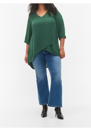 Femme portant une tunique asymétrique vert foncé, un jean évasé bleu et des bottines noires, debout sur un fond blanc.