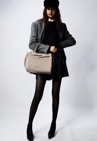 Beige leren handtas met gestructureerd ontwerp, bovenhandvatten, gouden hardware-accenten, leunend tegen een grijze blazer en een zwarte rok.
