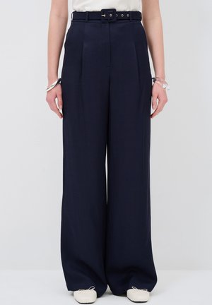 Frau trägt marineblaue Hosen mit weitem Bein und passendem Gürtel, weißes, eingestecktes Oberteil, weiße Ballerinas und durchsichtige Armbänder an beiden Handgelenken.