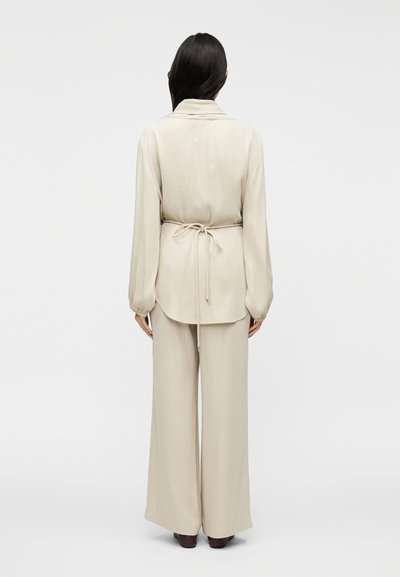 Μπεζ πουκάμισο με μακριά μανίκια, ζώνη στη μέση και πλισέ πίσω μέρος, συνδυασμένο με μπεζ παντελόνι φαρδιάς γραμμής. Λείο, ελαφρύ ύφασμα· μινιμαλιστικός σχεδιασμός.