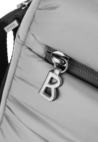 Angolo di uno zaino grigio imbottito con una zip in metallo che presenta la lettera "B". È visibile del materiale per le cinghie nero. Texture morbida in tutto il prodotto.