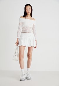 Top bianco in pizzo off-the-shoulder, gonna bianca a campana e sneaker argento con dettagli trasparenti; porta una piccola borsa bianca.