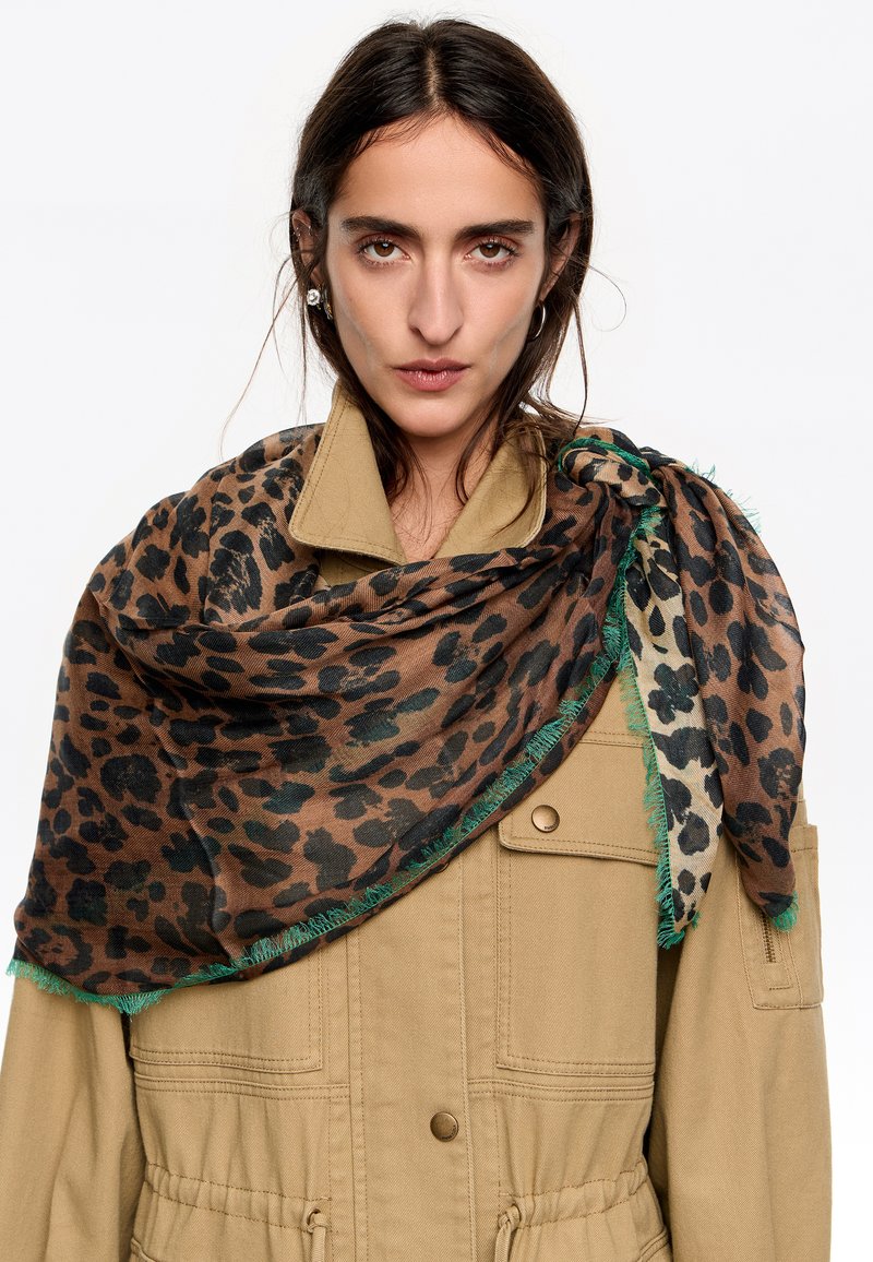 Bufanda de estampado de leopardo en marrón, con bordes deshilachados en verde, colocada sobre una chaqueta beige. La bufanda tiene un acabado suave y texturizado.