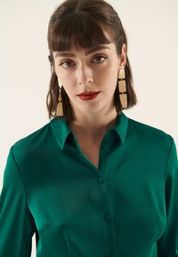 Blusa in raso verde smeraldo con chiusura a bottoni, maniche lunghe e colletto appuntito. Presenta una texture liscia e una lucentezza sottile.