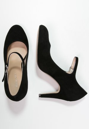 Chaussures à talons hauts en suède noir avec un bout rond, une sangle à la cheville avec des éléments en métal argenté, et une semelle intérieure beige lisse, présentant une silhouette élégante.