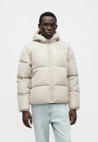 Svetlo béžová puffer bunda s kapucňou, s prešívaným dizajnom, predným zipsom a pruhovanými manžetami, spárovaná so svetlomodrými džínsami.