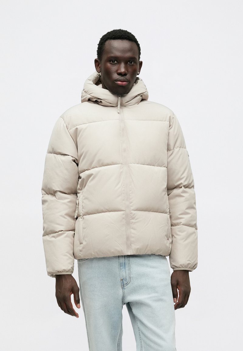 Svetlo béžová puffer bunda s kapucňou, s prešívaným dizajnom, predným zipsom a pruhovanými manžetami, spárovaná so svetlomodrými džínsami.