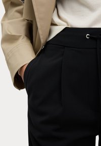 Beige jacka med en avslappnad passform, kombinerad med svarta plisserade byxor med sidofickor och dragsko i midjan. Texturerade tygdetaljer synliga.