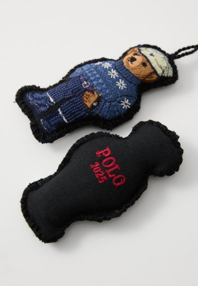 Izvezena plišana privjesak u obliku medvjeda u plavoj džemperici sa snjegovicama, uparen s crnom tkaninom oznakom "POLO 1925" u crvenoj boji.