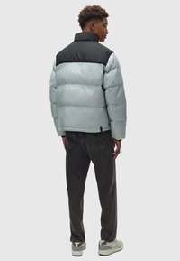 Hellblaues Puffermantel mit schwarzem oberen Abschnitt und hohem Kragen. Getragen mit dunklen Jeans und grauen Sneakers, was eine stromlinienförmige Silhouette zeigt.