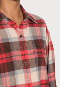 Chemise en flanelle à boutons avec motif à carreaux rouge, blanc, marron et bleu. Comprend un col, une poche poitrine et des boutons marron. Tissu doux et texturé.