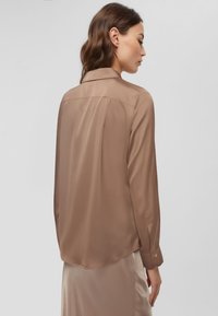 Blusa in satin marrone con colletto, maniche lunghe e polsini con bottone; texture liscia con vestibilità ampia e orlo arrotondato sul retro.