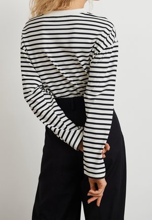 Haut à manches longues avec un motif à rayures horizontales noires et blanches, doté d'un col rond et de manches ajustées. Porté avec un pantalon noir.