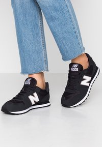 Svarta New Balance 500-sneakers med mesh- och syntetmaterial, framträdande vit "N"-logotyp och en texturerad sula. Bärs med ljusblå beskurna jeans.