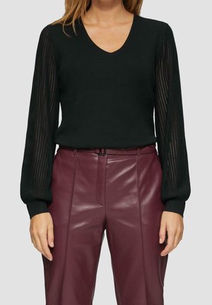 Femme portant un pull noir à col en V avec des manches longues à rayures transparentes et un pantalon en cuir bordeaux taille haute.