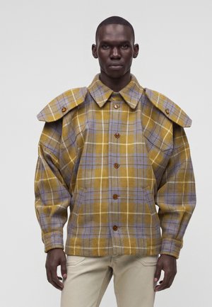 Homme portant une veste à carreaux jaune et violette boutonnée avec de grandes pattes d'épaule et un pantalon beige sur un fond uni.