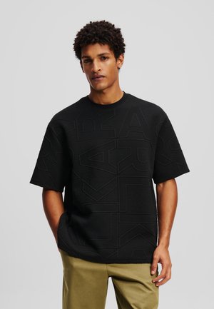 Black short-sleeve shirt with textured geometric patterns, round neckline, and relaxed fit, paired with olive green pants.