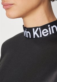 Czarny top z wysokim kołnierzem z białym logo "Calvin Klein". Materiał jest gładki o eleganckiej teksturze, dopasowany krój.