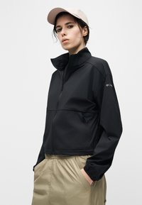 Νεαρό άτομο φοράει ανοιχτό ροζ καπέλο, μαύρο μπουφάν με φερμουάρ και μπεζ παντελόνι, στέκεται με τα χέρια στις τσέπες μπροστά σε απλό φόντο.