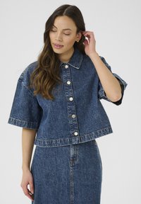 Nainen, jolla on päällä nappikiinnitteinen, lyhythihainen cropped denim-takki ja siihen sopivat korkeavyötäröiset denim-hameet, koskettaa hiuksiaan.