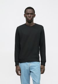 Mladý čierny muž v jednoduchom čiernom svetri s okrúhlym výstrihom a svetlomodrých džínsach, stojaci proti jednoliatej bielej stene.