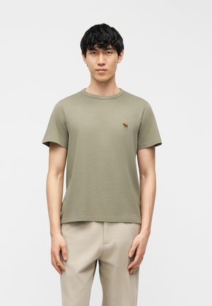 Mladić s crnom kosom koji nosi tamnozelenu majicu kratkih rukava s malim smeđim logom losa i bež hlače, stoji neutralno.