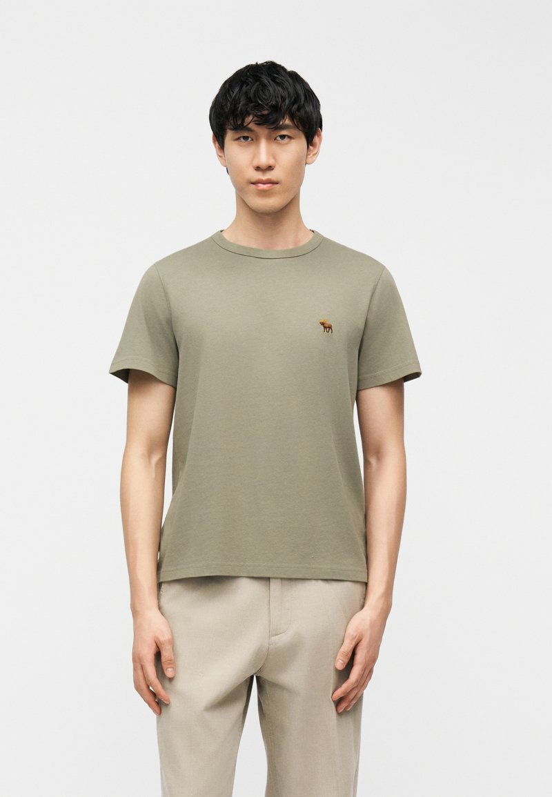 Νεαρός άνδρας με μαύρα μαλλιά, φοράει μια μουντή πράσινη κοντομάνικη μπλούζα με ένα μικρό καφέ λογότυπο με έναν ταράνδο και μπεζ παντελόνι, στέκεται ουδέτερα.