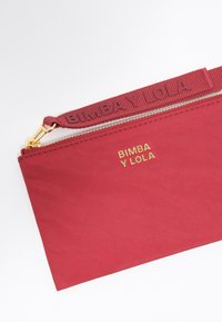 Cartera de nailon roja con el logotipo dorado "BIMBA Y LOLA", una correa texturizada y cremallera en tono dorado. Diseño simple, rectangular con un acabado liso.