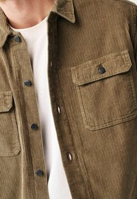 Braunes Cordhemd mit durchgehender Knopfleiste, zwei Brusttaschen und schwarzen Knöpfen; darüber ein weißes Hemd getragen. Strukturierte, gerippte Musterung.