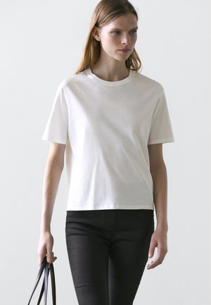 Frau mit langen Haaren, die ein schlichtes weißes kurzärmeliges T-Shirt und schwarze Hosen trägt, hält eine schwarze Handtasche mit dünnen Tragegurten.