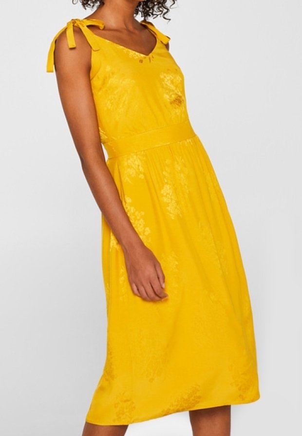 Femme portant une robe midi jaune sans manches avec des bretelles nouées aux épaules et des motifs floraux subtils.