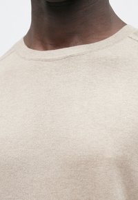 Bež pulover iz pletenine z okroglim izrezom ovratnika, mehko teksturo in prilegajočim krojem. Vidni šivi na ovratniku dodajajo subtilne podrobnosti.