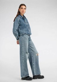 Giacca di jeans azzurro chiaro con un design corto e tasca frontale, abbinata a jeans azzurro chiaro a gamba larga con aree distressed e vita alta.