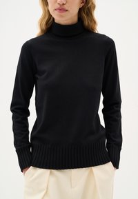 Maglione a collo alto nero realizzato in morbido tessuto a maglia. Caratterizzato da maniche lunghe e orlo a costine. Abbinato a pantaloni beige chiaro.