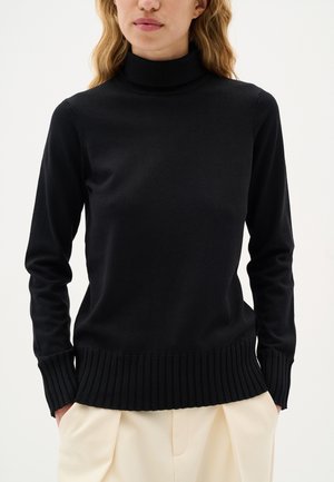 Schwarzer Rollkragenpullover aus weichem Strickmaterial. Mit langen Ärmeln und einem gerippten Saum. Kombiniert mit hellbeigen Hosen.