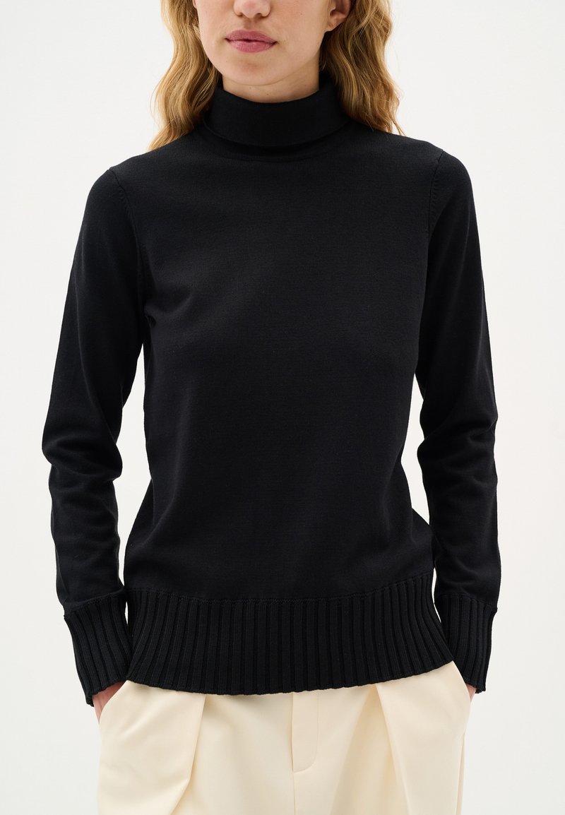 Maglione a collo alto nero realizzato in morbido tessuto a maglia. Caratterizzato da maniche lunghe e orlo a costine. Abbinato a pantaloni beige chiaro.