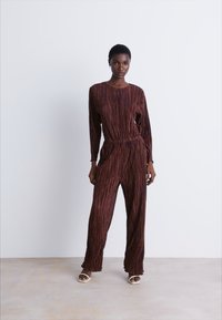 Combinaison rayée marron avec des manches longues et une taille élastique. Le modèle porte des chaussures à bout ouvert. Texture lisse et coupe décontractée.