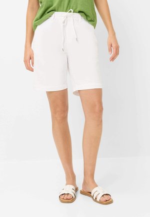Person trägt weiße Shorts mit Kordelzug, grünes Oberteil und weiße flache Sandalen, steht vor weißem Hintergrund.