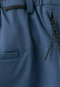 Tissu bleu avec une poche zippée, cordon de serrage noir avec des accents blancs, et une étiquette noire "MNG PERFORMANCE" près de la taille.