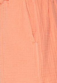 Pantalones de tela texturizada color durazno claro con bolsillos laterales y costuras visibles. El material tiene un aspecto arrugado, ofreciendo un ajuste relajado.