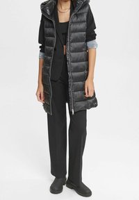 Musta puffervesti kiiltävällä pinnalla, vetoketjullinen etuosa ja ylisuuri huppu, yhdistettynä mustiin leveisiin housuihin ja nilkkasaappaisiin.