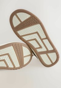 Sulan på en sko med en vit gummibas och ett brunt texturerat mönster samt geometriska former, designad för grepp och stabilitet.