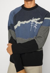 Homme portant un pull à manches longues en maille avec des blocs de couleur noir, bleu et gris, et des motifs d'arbres blancs sur la poitrine et la manche.